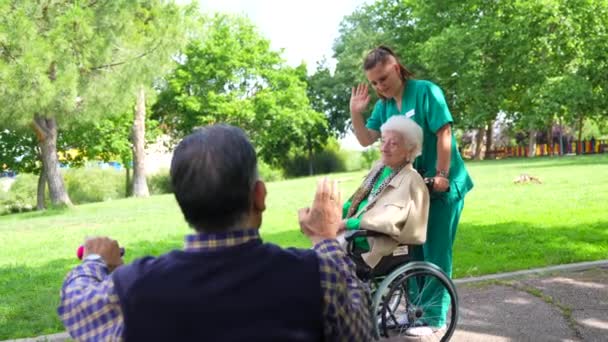 一位老年妇女带着护士步行穿过疗养院的花园 坐在轮椅上迎接一位老年男子 — 图库视频影像