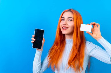Stüdyo fotoğrafçılığındaki kızıl saçlı kadın mavi arka planda cep telefonu ile online ödeme yapıyor.