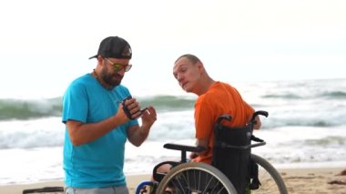 Plajda tekerlekli sandalyede sakat bir adam ve bir arkadaşı selfie çekiyor.