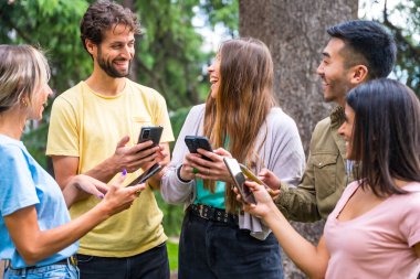 Parkta internet ya da sosyal ağları izleyen çok ırklı gülümseyen bir grup, teknoloji kavramı