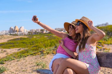 Şapkalı kız arkadaşlar plaj tatilinde çok mutlular. Telefonla selfie çekiyorlar. Yaz tatili arkadaşlarıyla arkadaşlık konsepti.