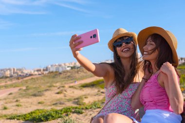 Plajda şapkalı bir kadın telefonda gülümserken selfie çekiyor.