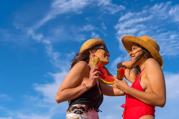 stock image Female friends at the beach in summer smiling eating a watermelon with the sky in the background, bottom up shot