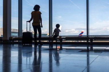 Havaalanının penceresinin yanında bavul taşıyan anne ve oğlunun silueti uçaklara bakıyor.