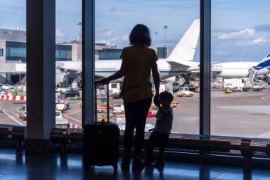 Havaalanının penceresinin yanında duran valizli anne-oğul silueti, tatil kavramı.