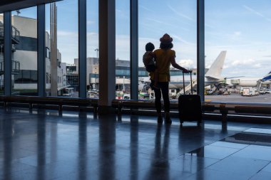 Anne ve oğlu pencerenin yanında bavullarıyla havaalanındaki uçaklara bakıyorlar.