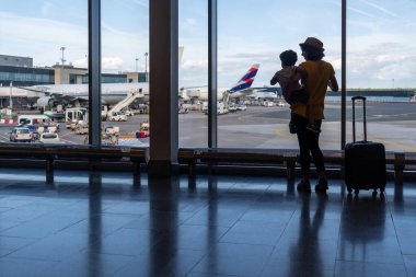 Anne ve oğlu pencerenin yanında bavullarıyla havaalanındaki uçaklara bakıyorlar.