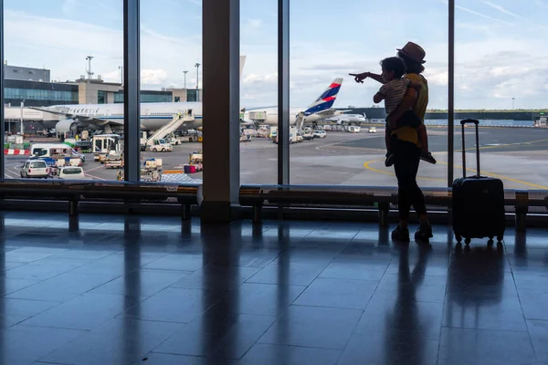 Anne ve oğlu pencerenin yanında bavullarıyla havaalanı uçaklarına bakıyor, yaz tatili konsepti.