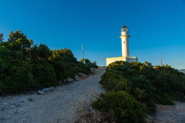 Lefkada adasının güney bölgesinde gün batımında beyaz deniz fenerine veya Cape Ducato Lefkas Burnu 'na giden yol. Yunanistan.