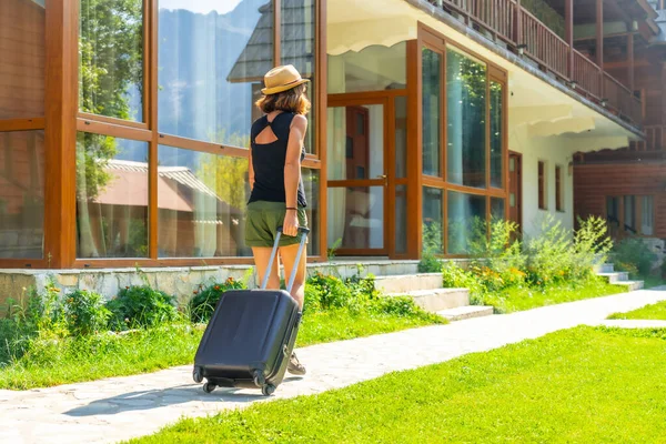 Tourist woman with a suitcase and hat on rural vacation arriving at a mountain cabin, summer vacation in nature