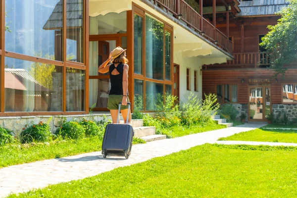 Tourist woman with a suitcase and hat arriving at a mountain cabin, summer rural vacation in nature