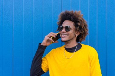 Cep telefonuyla konuşan Afro-Amerikan bir gencin mavi arka planlı yatay fotoğrafı.