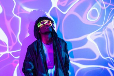 Studio portrait with purple and blue neon lights of an afro futuristic man looking up using augmented reality goggles clipart