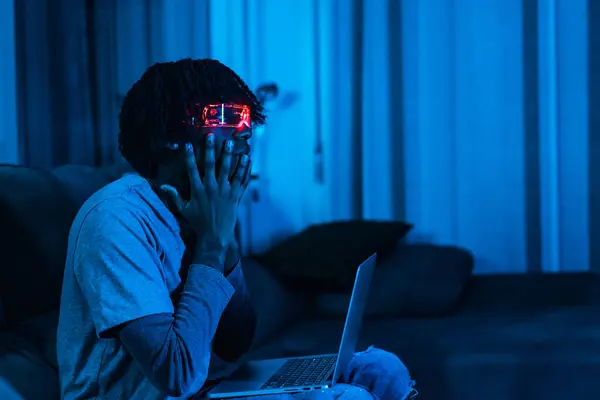 Afro man using laptop and Augmented reality goggles in a living room with neon blue light