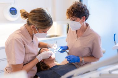 Bir kliniğin sandalyesinde yatan bir hastanın ağzını inceleyen dişçi ve asistan.