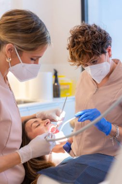 Bir dişçi kliniğinde sandalyeye yaslanmış bir kadının ağzını inceleyen teknisyen.