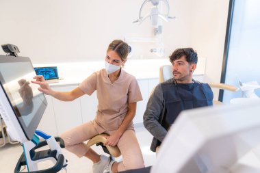Modern bir klinikte bir sandalyede oturan hastaya çok boyutlu görüntü gösteren dişçi.