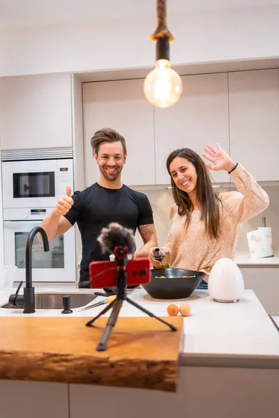 Vertical photo of a team of vloggers waving at camera while making video cooking in a domestic kitchen