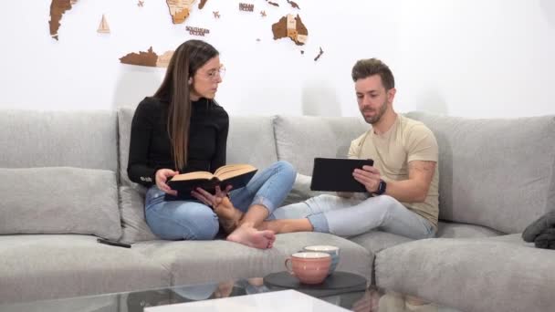 Wanita Membaca Sebuah Buku Samping Pasangannya Menggunakan Tablet Yang Duduk — Stok Video