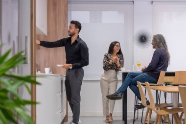 Üç iş arkadaşı kahve molası sırasında iş yerinin mutfağında rahatça konuşuyorlar.