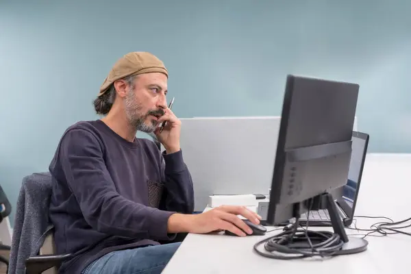 Modern and stylish mature man talking to the phone while using computer in a co-working