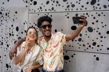 Kentsel bir arka planda cep telefonuyla selfie çekerken çok ırklı arkadaşlar kucaklaşıyor