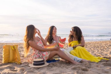 Üç çok ırklı arkadaşın plajda karpuz yerken eğlendiği huzurlu yaz pikniği.