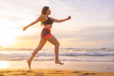 Gün batımında sahilde koşan güçlü fit bir kadının yan görüntüsünün olduğu yatay fotoğraf.