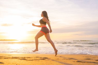 Gün batımında sahilde koşan genç bir kadının yan görüntüsünün olduğu yatay fotoğraf.