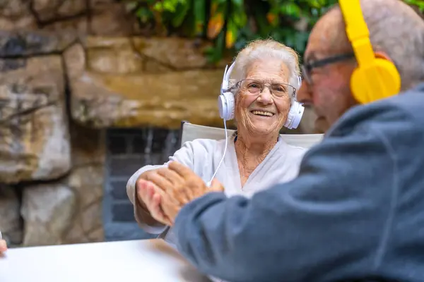 Happy elder people using headphones and smiling sitting in a garden in a geriatric