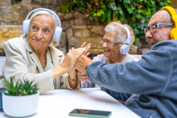 Пожилые люди наслаждаются музыкой, используя наушники, сидя в саду в доме престарелых