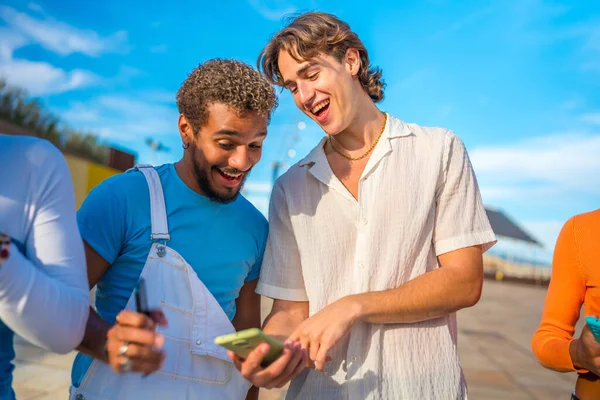 Smiling and distracted multi-ethnic young men using phone outdoors next to friends