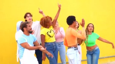 Farklı etnik gruplardan oluşan bir grup sokak ortasında, sarı bir arka planın yanında ortak bir koreografi gösterisi yapıyor.