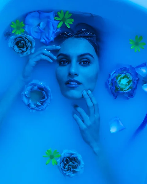 Artistic blue colored portrait from above of a sensual woman in a bath with milk and flowers