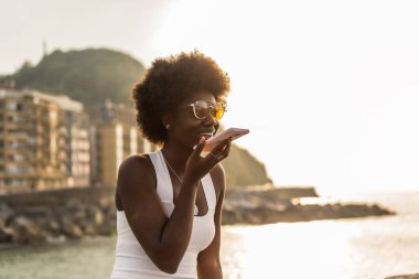 Mutlu, genç bir Afrikalı kadının gün batımında deniz kenarında sesli mesaj gönderirken yan görüntüsü.