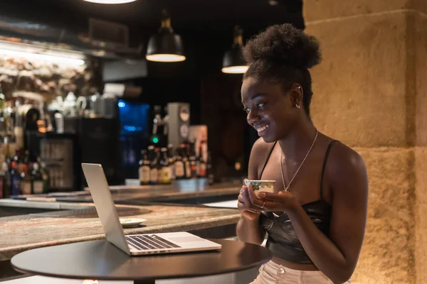 Side view of a happy and young relaxed woman sitting on a bar during online meeting