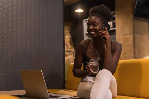 Cheerful african young woman talking to the mobile working on remote sitting on a bar