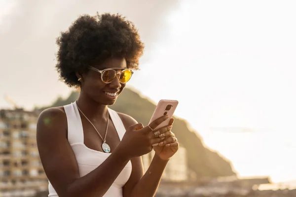 Happy trendy african young woman using phone outdoors during sunset next to the sea