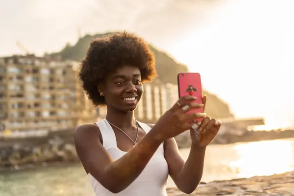 Happy afro woman in white dress taking a selfie during sunset next to the sea