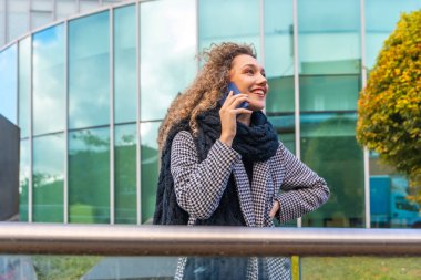 Sıcak kıyafetler içinde gülümseyen kadın sonbaharda şehir caddesinde cep telefonuyla konuşuyor.