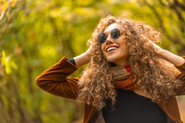 Kıvırcık saçlı, güneş gözlüklü, sonbaharda parkta poz verip gülümseyen mutlu bir manken.