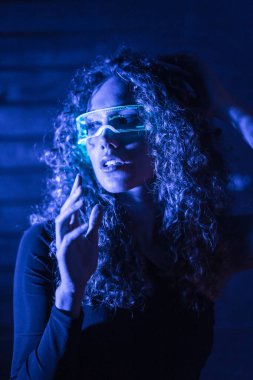 Koyu mavi bir alanda akıllı gözlük kullanan şehvetli bir kadının dikey portresi.