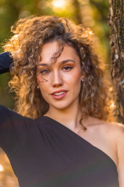 Ormandaki kıvırcık saçlı ve şehvetli elbiseli genç bir mankenin dikey yakın çekimi.
