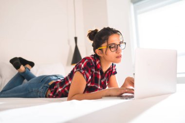 Otel yatağında dizüstü bilgisayarla çalışan genç bir kadının yan görüntüsü.