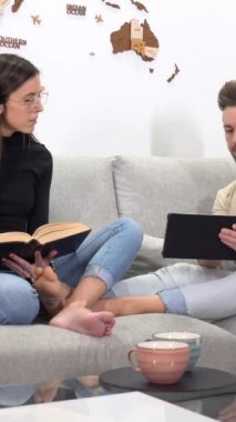 Yanında kitap okuyan bir kadın evdeki kanepede birlikte oturup tablet kullanıyor.