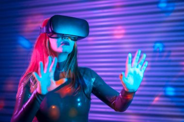 Sarışın bir kadın, interaktif bir oyun sırasında kentsel bir gece mekanında neon ışıklı VR gözlükleriyle...