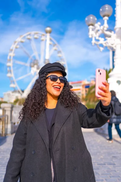 Vertical photo of a latin chic woman taking a selfie walking along a city in a sunny day of winter