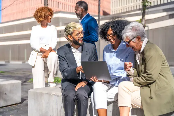 Multi-ethnic business people using laptop sitting outdoors next to a financial building