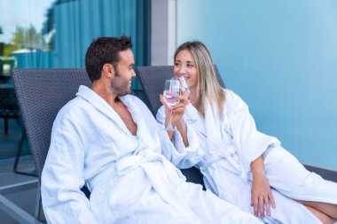 Lüks bir otelde beyaz bornoz giymiş beyaz şarapla kadeh kaldırarak hayal kuran çift.