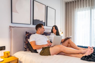 Rahatlamış bir adamın sabah sahnesinde karısının yanında dizüstü bilgisayar kullanarak yatakta kitap okuması.